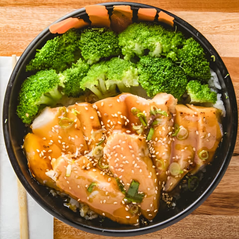 Teriyaki chicken with sesame seeds, green onions, and broccoli over rice in a black bowl.